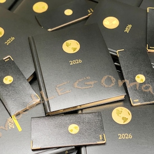 May include: A collection of black 2026 planners and notebooks. Each has a gold foil world map emblem and the year 2026 printed in gold. Some have gold page edges and a gold corner. The items are in various sizes.