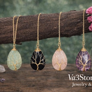 May include: Four gemstone pendant necklaces with gold chains, each featuring a tree of life design. The gemstones are light green, black, pink, and purple. The necklaces are displayed on a wooden branch.