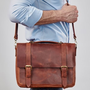 May include: A brown leather messenger bag with a long adjustable strap. The bag has two buckles on the front and the initials "LMT" are embossed on the front flap.