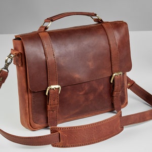 May include: A brown leather satchel bag with a long adjustable shoulder strap and a top handle. The bag has two buckles on the front and a flap closure.
