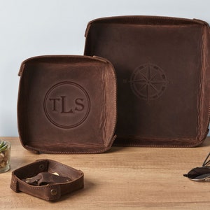 May include: Two brown leather trays with a compass design and the initials "TLS" embossed on one of the trays. The trays are on a wooden surface with a small plant and sunglasses.
