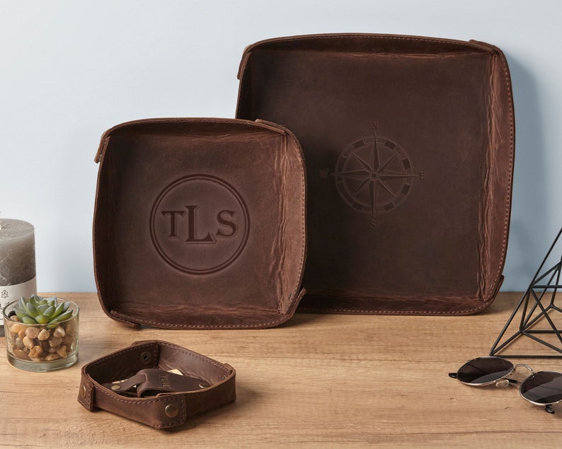 May include: Two brown leather trays with a compass design and the initials "TLS" embossed on one of the trays. The trays are on a wooden surface with a small plant and sunglasses.