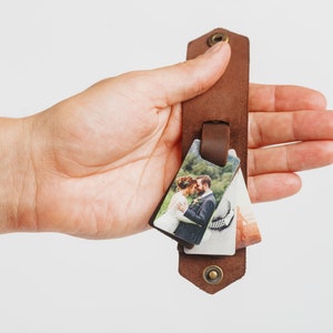 Puede incluir: Una pulsera de cuero marr&oacute;n con tres peque&ntilde;os colgantes de fotos rectangulares. Las fotos representan escenas de boda. La pulsera tiene un cierre de bot&oacute;n a presi&oacute;n y un detalle de remache met&aacute;lico.