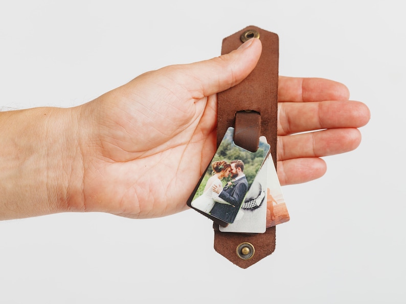 Puede incluir: Una pulsera de cuero marr&oacute;n con tres peque&ntilde;os colgantes de fotos rectangulares. Las fotos representan escenas de boda. La pulsera tiene un cierre de bot&oacute;n a presi&oacute;n y un detalle de remache met&aacute;lico.