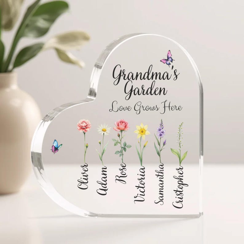 May include: A clear heart-shaped acrylic plaque with the text "Grandma's Garden Love Grows Here" and a row of flowers with names below each flower: Oliver, Adam, Rose, Victoria, Samantha, Christopher.  There are butterflies on the plaque.