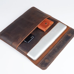 May include: A brown leather laptop sleeve with a flap that opens to reveal a pocket for a phone and a separate pocket for a wallet. The sleeve is open and a laptop is inside.