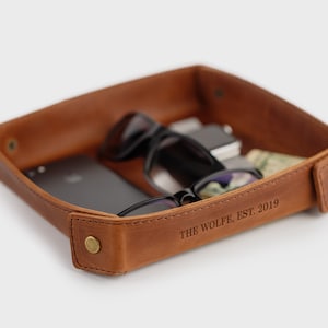 May include: A brown leather tray with a personalized inscription that reads "THE WOLFE, EST. 2019". The tray is filled with various items, including a smartphone, sunglasses, and a wallet.