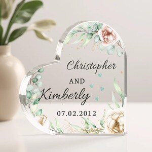 May include: A clear heart-shaped acrylic keepsake with a floral design and the text "Christopher and Kimberly 07.02.2012".