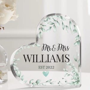 May include: A clear heart-shaped acrylic plaque with a leafy design and the text "Mr. & Mrs. WILLIAMS EST 2022" with a small teal heart.