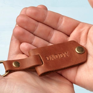 Puede incluir: Un llavero de cuero marr&oacute;n con la palabra "DADDY" grabada en relieve. El llavero tiene un anillo y remaches de metal color lat&oacute;n. El cuero es de un color marr&oacute;n medio y c&aacute;lido.