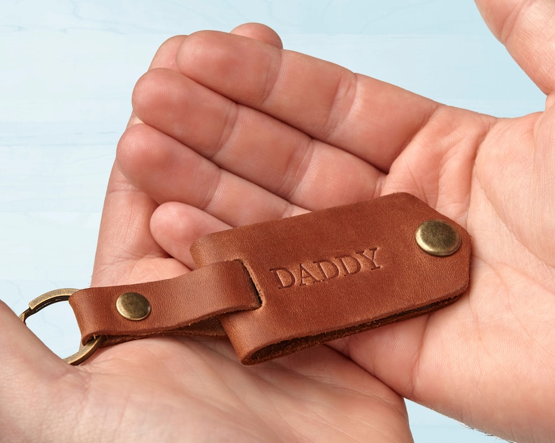 Puede incluir: Un llavero de cuero marr&oacute;n con la palabra "DADDY" grabada en relieve. El llavero tiene un anillo y remaches de metal color lat&oacute;n. El cuero es de un color marr&oacute;n medio y c&aacute;lido.