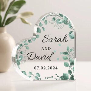 May include: A clear heart-shaped acrylic keepsake with a leafy green design and the text "Sarah and David 07.02.2024".