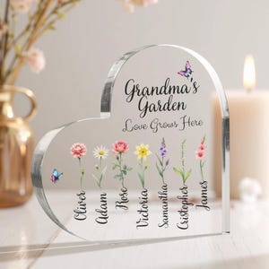 May include: A clear heart-shaped acrylic plaque with the text "Grandma's Garden Love Grows Here" and a butterfly design.  The plaque features seven flowers with names written below each flower: Oliver, Adam, Rose, Victoria, Samantha, Christopher, and James.