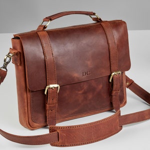 May include: A brown leather briefcase with a top handle and a shoulder strap. The bag has two buckles on the front and the initials "DC" are embossed on the front.
