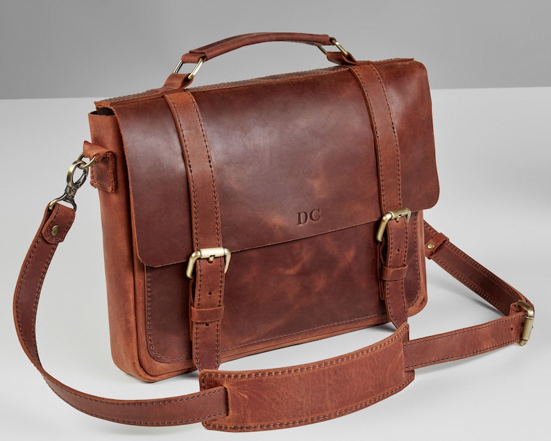 May include: A brown leather briefcase with a top handle and a shoulder strap. The bag has two buckles on the front and the initials "DC" are embossed on the front.
