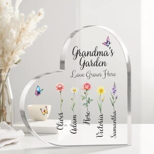 May include: A clear heart-shaped acrylic plaque with the text "Grandma's Garden Love Grows Here". Five flowers with names "Oliver", "Adam", "Rose", "Victoria", and "Samantha" are displayed below the text. Butterflies are also included in the design.