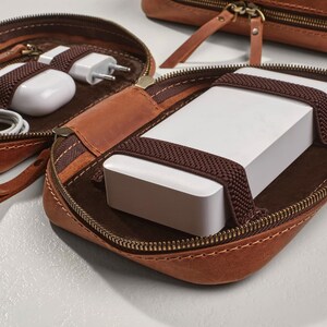 以下が含まれることがあります： ジッパー付きの茶色の革製のトラベルケース。ケースが開いており、白いモバイルバッテリーが入っています。茶色のゴムバンドで固定されています。