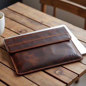 May include: A brown leather laptop sleeve with a flap closure. The sleeve has a distressed finish and is personalized with the initials "PAM". The sleeve is resting on a wooden table.