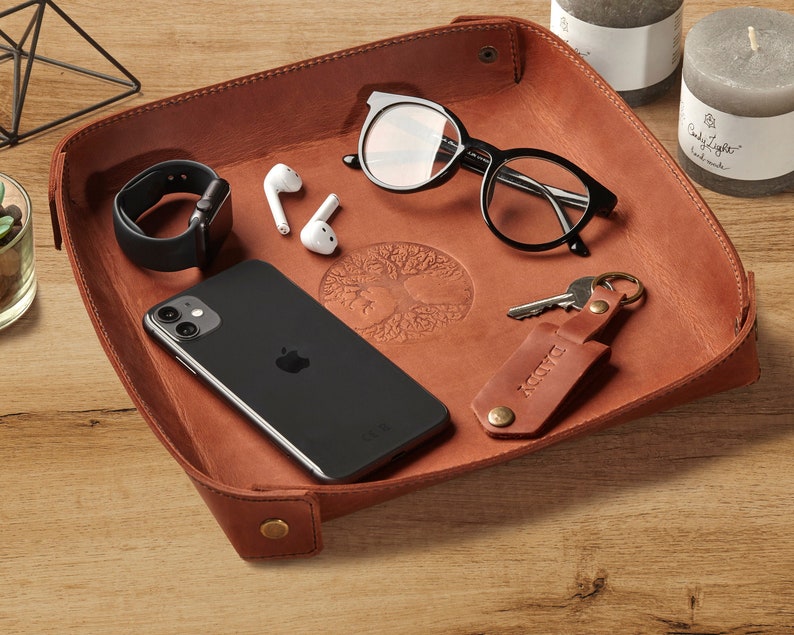 May include: A brown leather tray with a tree design embossed in the center. The tray contains a smartphone, a watch, a pair of glasses, a set of keys with a leather keychain that says "DADDY", and a pair of wireless earbuds.