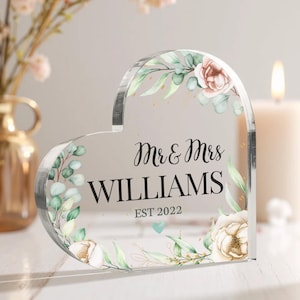 May include: Clear heart-shaped acrylic wedding sign with a floral design and the text "Mr & Mrs WILLIAMS EST 2022".