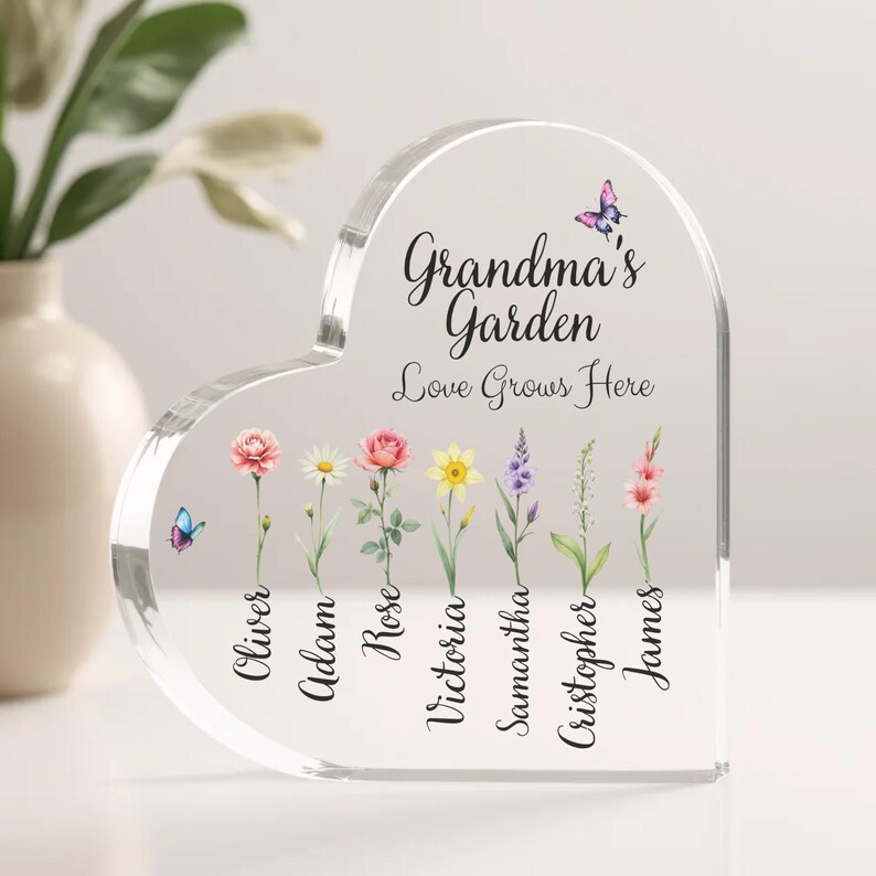 May include: A clear heart-shaped acrylic plaque with the text "Grandma's Garden Love Grows Here" and a butterfly design. Seven flowers with names written below them: Oliver, Adam, Rose, Victoria, Samantha, Christopher, and James.