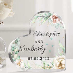 May include: A clear heart-shaped acrylic keepsake with a floral design and the text "Christopher and Kimberly 07.02.2012".
