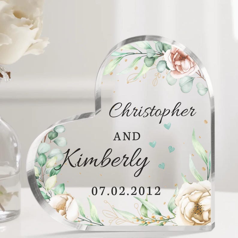 May include: A clear heart-shaped acrylic keepsake with a floral design and the text "Christopher and Kimberly 07.02.2012".
