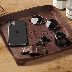 May include: A brown leather valet tray with a personalized monogram "TLS" in a circle. The tray contains a smartphone, sunglasses, wireless earbuds, a watch, and keys.