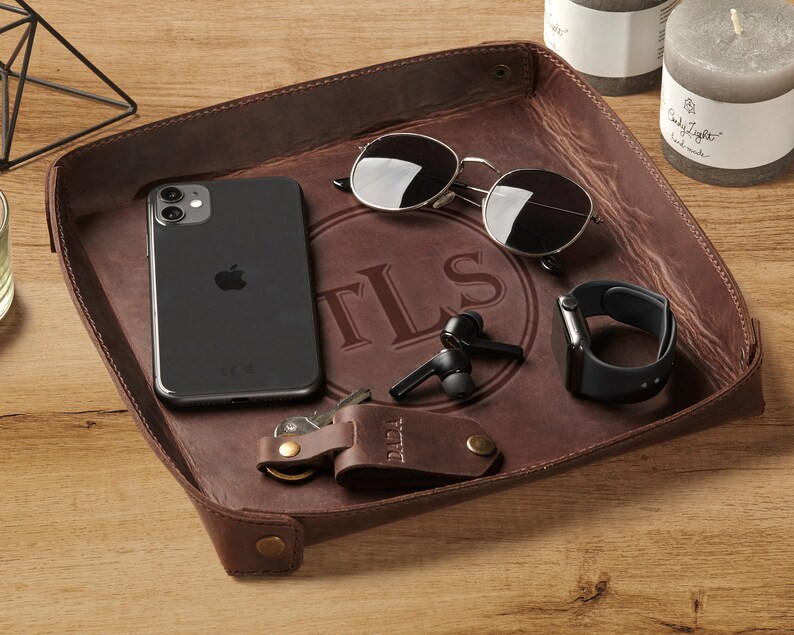 May include: A brown leather valet tray with a personalized monogram "TLS" in a circle. The tray contains a smartphone, sunglasses, wireless earbuds, a watch, and keys.