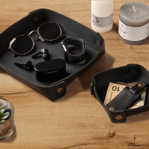 May include: Two black leather trays with rounded corners. The larger tray contains sunglasses, a pair of wireless earbuds, a small black case, and a black smartwatch. The smaller tray contains a key chain with a leather tag that says "Daddy" and a small piece of paper with the number "10" written on it.