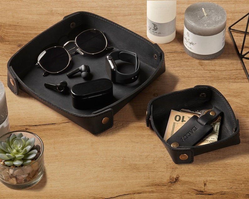 May include: Two black leather trays with rounded corners. The larger tray contains sunglasses, a pair of wireless earbuds, a small black case, and a black smartwatch. The smaller tray contains a key chain with a leather tag that says "Daddy" and a small piece of paper with the number "10" written on it.