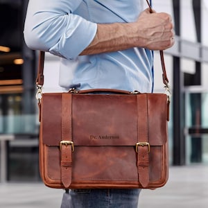 May include: A brown leather satchel with a shoulder strap and gold-colored buckles. The bag has a flap closure and a handle on top. The text "Dr. Anderson" is engraved on the front of the bag.