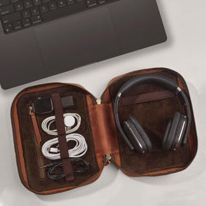 May include: A brown leather travel case with a zipper closure. The case is open and contains a pair of black headphones, a watch, and several cords.