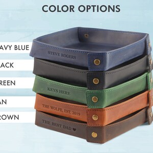May include: Five leather trays stacked on top of each other. The trays are in the colors navy blue, black, green, tan, and brown. Each tray has a different personalized inscription.