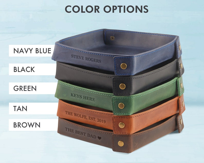 May include: Five leather trays stacked on top of each other. The trays are in the colors navy blue, black, green, tan, and brown. Each tray has a different personalized inscription.