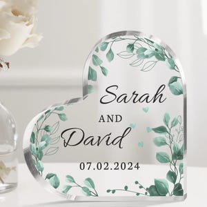 May include: A clear heart-shaped acrylic wedding keepsake with a leafy green floral design. The text reads "Sarah and David 07.02.2024".