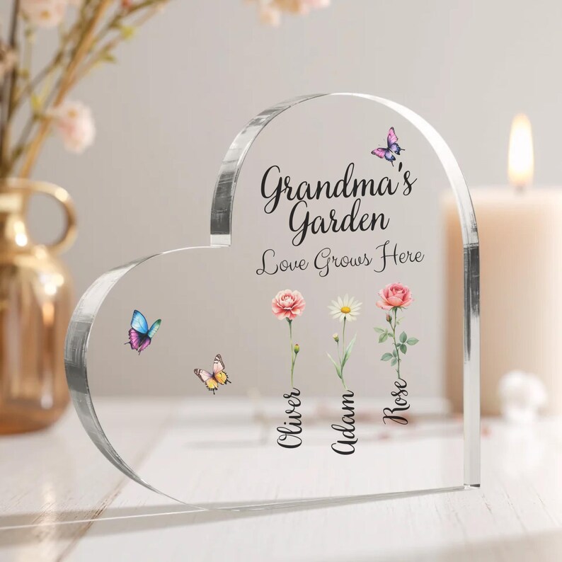 May include: A clear heart-shaped acrylic plaque with the text "Grandma's Garden Love Grows Here" and three flowers with the names "Oliver", "Adam", and "Rose" written below them. There are butterflies on the plaque.