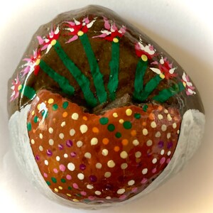 May include: A painted rock with a brown background and a white rim. The rock is painted to look like a flower pot with pink and white flowers and green stems. The pot is filled with brown, orange, yellow, green, white, and purple dots.