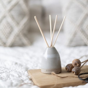 May include: A grey ceramic diffuser bottle with wooden sticks and the word "Soomish" on the bottom. The diffuser is sitting on a wooden tray with dried poppy seed pods.