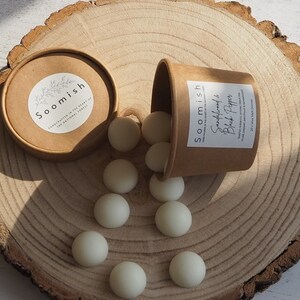 May include: A brown paper container with white wax melts labelled "Soomish's Sandalwood & Black Pepper" on a wooden surface.