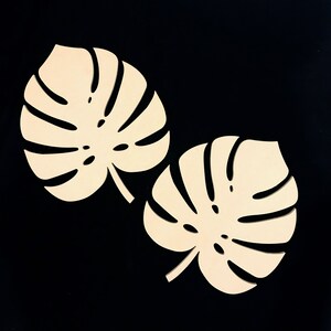 Puede incluir: Dos recortes de madera de color beige claro, planos, con forma de hojas de monstera. Cada hoja presenta las caracter&iacute;sticas hendiduras profundas y alargadas junto con perforaciones centrales, mostrando una superficie lisa y mate.