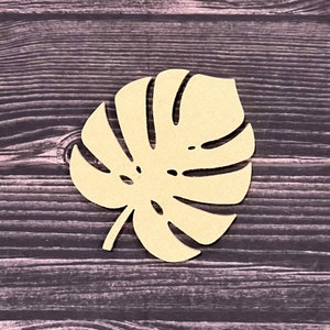 Puede incluir: Un recorte de madera de color beige claro, plano y cortado con l&aacute;ser, con forma de hoja de monstera. La hoja presenta l&oacute;bulos profundos caracter&iacute;sticos y varias perforaciones ovaladas en toda su superficie, imitando el aspecto natural.