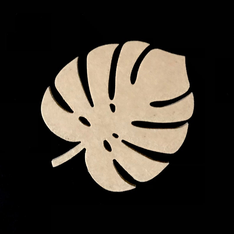 Puede incluir: Un recorte de madera de color beige claro con forma de hoja de monstera. El objeto presenta un dise&ntilde;o distintivo y sim&eacute;trico con varios recortes alargados y curvos que irradian desde el centro, imitando la nervadura natural de la planta.