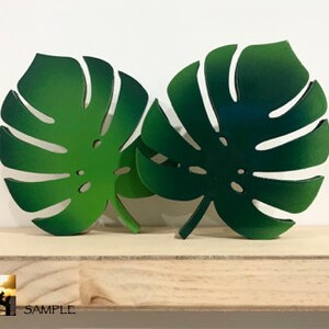 Puede incluir: Dos recortes decorativos en forma de hoja de monstera que presentan un degradado de tonos verdes, desde el m&aacute;s claro al m&aacute;s oscuro. Cada hoja muestra el dise&ntilde;o caracter&iacute;stico de la planta monstera con sus divisiones y perforaciones.