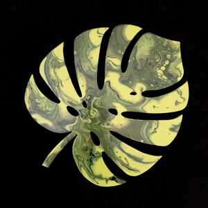 Puede incluir: Un recorte con forma de hoja de monstera que presenta un dise&ntilde;o de pintura vertida marmolada en tonos amarillo p&aacute;lido, verde salvia y verde bosque oscuro. La superficie muestra patrones fluidos y arremolinados de colores mezclados.
