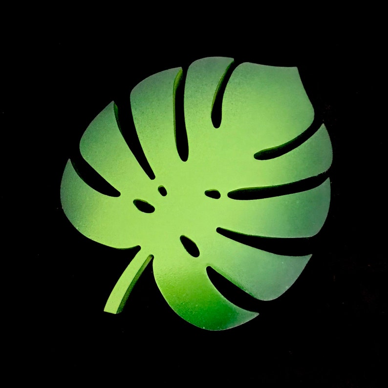 Puede incluir: Un posavasos con forma de hoja de monstera de color verde brillante con un acabado mate y suave. La forma de la hoja presenta recortes profundos caracter&iacute;sticos y peque&ntilde;as perforaciones interiores, creando una silueta distintiva.
