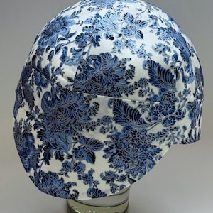 Könnte beinhalten: Schweißkappe mit weißem und blauem Blumenmuster. Die Kappe zeigt ein sich wiederholendes Design aus blauen Blumen und Blättern auf weißem Grund. Sie schützt Kopf und Haar vor Funken und Hitze.