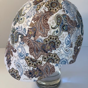 May include: A patterned newsboy cap featuring a paisley design in shades of brown, blue, and green on a white background. The cap is displayed on a clear glass object, showcasing its shape and design. The cap is a stylish accessory.
