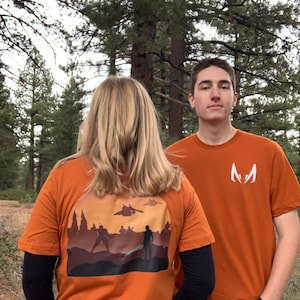 May include: Two people wearing orange t-shirts with different designs. The person on the left is wearing a t-shirt with a sunset scene and the text "M" on the back. The person on the right is wearing a t-shirt with a white "M" on the front.