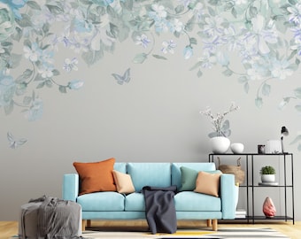 May include: A light blue sofa with orange, beige, and green throw pillows and a gray throw blanket. The sofa is in front of a gray wall with a floral wallpaper border. There is a black metal shelf with a potted plant and a lamp on the right side of the image.